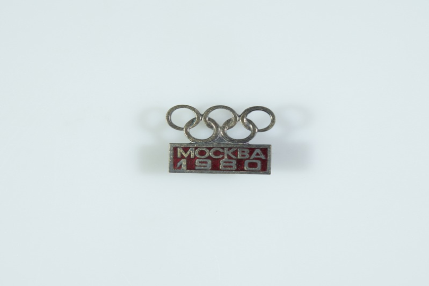Eine weiße Fläche mit einem Abzeichen mit der Aufschrift "Mockba 1980 Olympische Spiele" und einer Nadel mit den Olympischen Ringen.