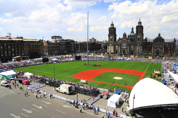 Große Menschenmenge auf einem Fußballfeld in Mexiko-Stadt mit Gebäuden, Bäumen, Laternenpfählen, Fahrzeugen und einem Zelt im Hintergrund unter einem bewölkten Himmel.