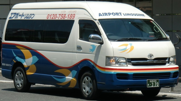 Weißer und blauer Van am Straßenrand mit einem Gebäude im Hintergrund, das ein sichtbares Vorderrad zeigt.