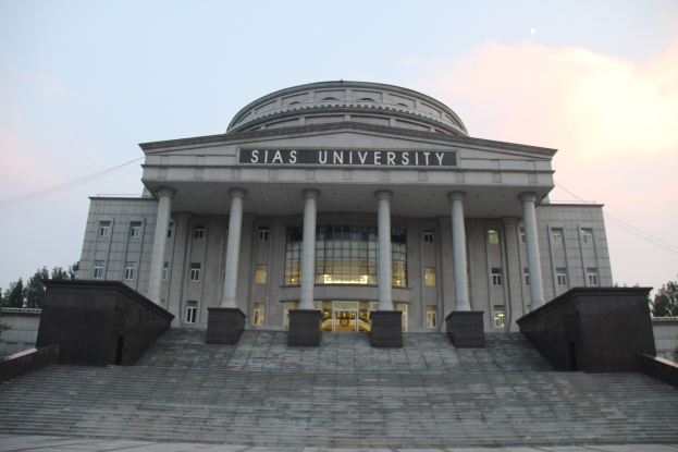 Large building with pillars, windows, doors, and steps, surrounded by trees under a clear blue sky, with the university's name displayed on the facade.