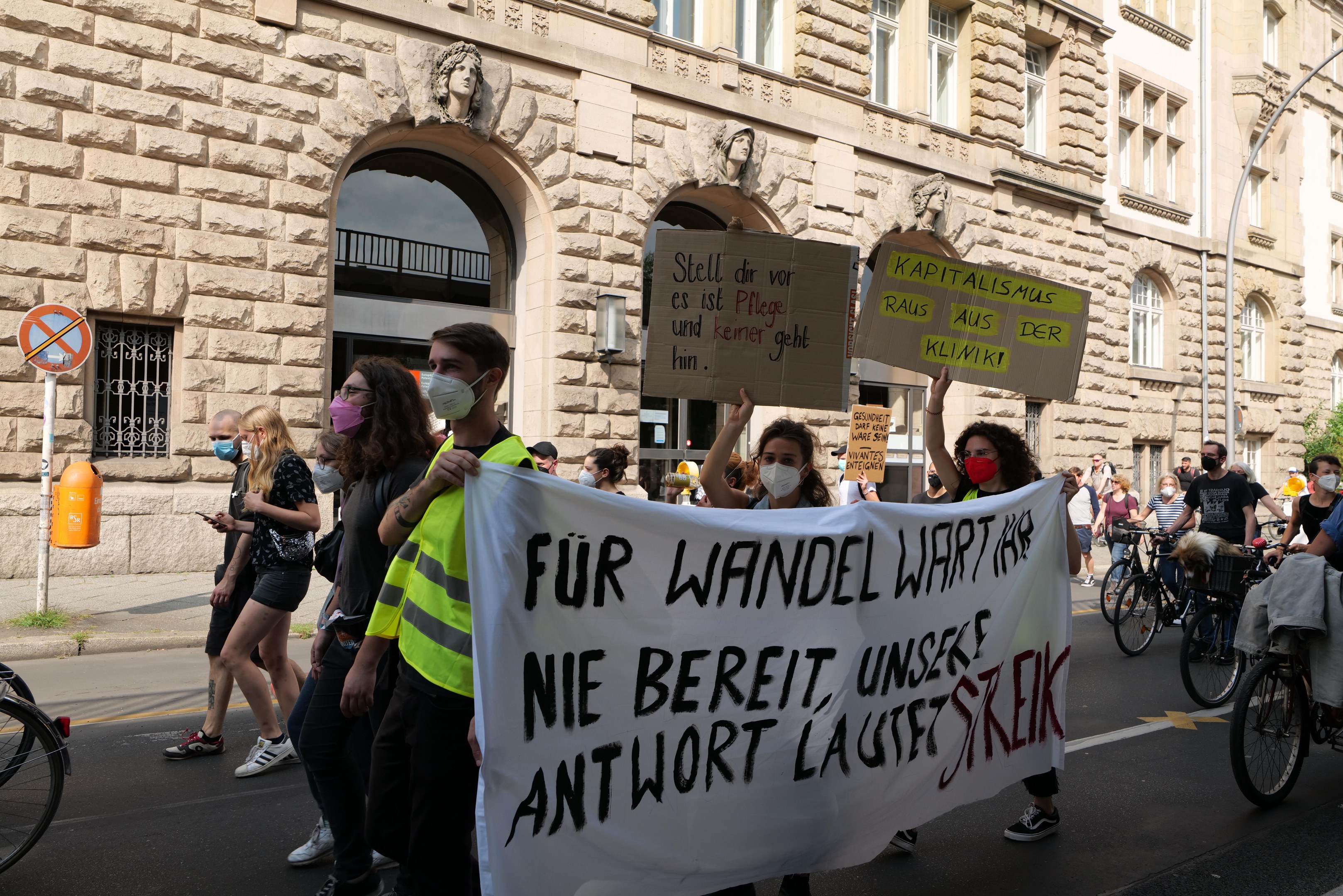 A group of people marching in a protest on a city street, some holding signs and others riding bicycles, with a historic building featuring windows, arches, pillars, and sculptures in the background.