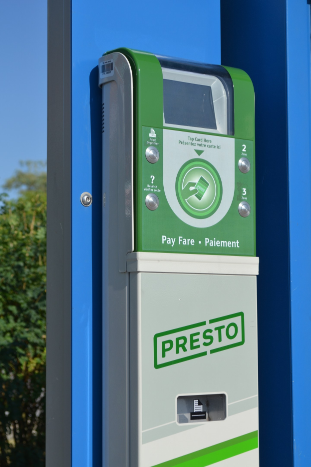 Ein grüner und weißer Parkscheinautomat mit Knöpfen und Text auf einer blauen Wand, mit Bäumen und einem klaren blauen Himmel im Hintergrund.