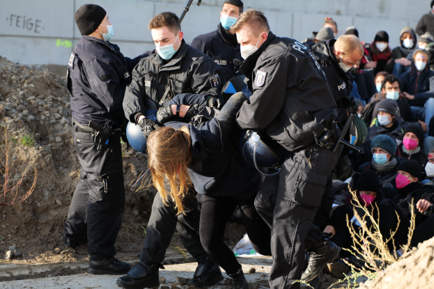 Gruppe maskierter Personen in Mützen, einige bewaffnet, um eine Frau herum, die von der Polizei begleitet wird, mit städtischen Hintergrund-Elementen wie Pflanzen, Steinen und einer Wand mit textbezogenen Protesten.