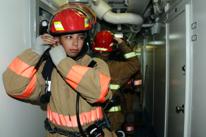 Eine Gruppe von Feuerwehrleuten in Uniform steht zusammen in einem Raum mit einer Tür auf der rechten Seite und einer Wand auf der linken Seite, mit Rohren und anderen Gegenständen im Hintergrund, was auf eine Übung hindeutet.