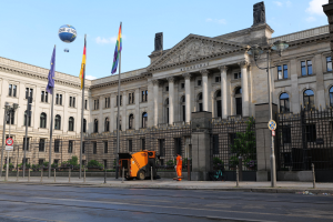 Large modern government building with many windows, surrounded by street furniture and vehicles, with the German Bundestag flag visible.