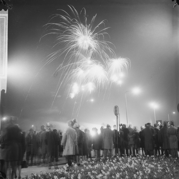 Eine Menschenmenge steht nachts vor einem Gebäude, das von Straßenlaternen beleuchtet wird und von blühenden Pflanzen umgeben ist, während farbige Feuerwerke den Himmel erhellen.