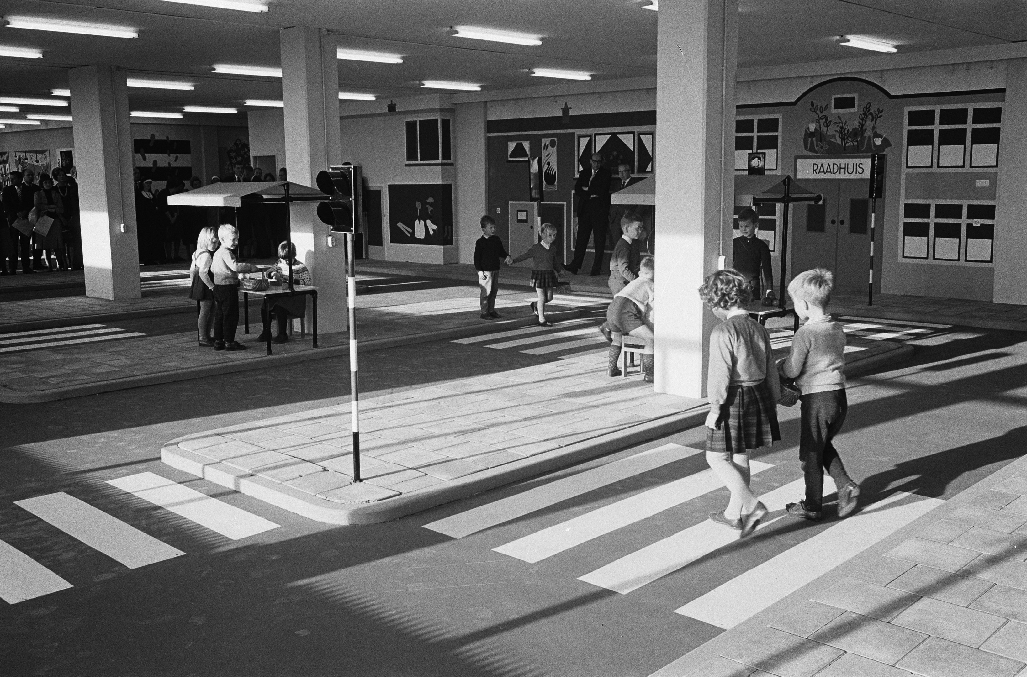 Schwarz-weiß-Foto von Kindern, die eine Straße an einer Ampel überqueren, mit Fußgängern in der Nähe, einem Verkehrssignalmast im Vordergrund und Hintergrundsäulen mit Deckenleuchten.