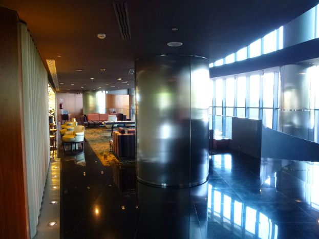 Eine geräumige Business-Lounge am Hong Kong International Airport mit großen Fenstern, hohen Decken mit elektrischen Leuchten, Sitzgruppen einschließlich Stühle und Sofas, Tischen, einem Teppichboden und einer zentralen Säule.