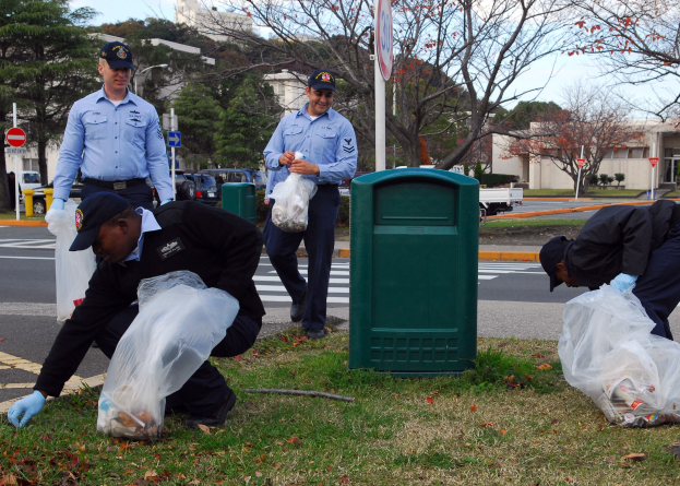 Drei Polizisten in Uniform sammeln Müll an einer Straßenrand mit Bäumen, Gebäuden, Fahrzeugen und einem Mülleimer im Hintergrund.