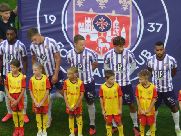 Eine Gruppe junger Menschen, die auf einem Fußballfeld zusammenstehen, mit einem Banner im Hintergrund, auf dem "Preston Primary School Football Team" steht.
