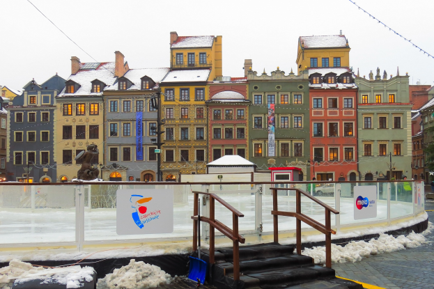 Eislaufbahn in einem von Gebäuden umgebenen Stadtplatz mit Treppen, Geländern, Informationsschildern, schneebedecktem Boden, Laternen, Bäumen und einem Himmel als Hintergrund.