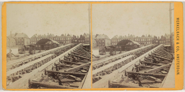 Ein altes Schwarz-Weiß-Foto einer Bahnschiene mit Gebäuden im Hintergrund, auf dem rechts der Text 'Merkelbach & Co. Amsterdam' steht und im Vordergrund verstreute hölzerne Gegenstände zu sehen sind.