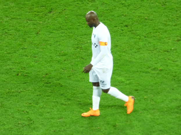 Ein Fußballspieler in einem weißen T-Shirt, weißen Shorts und orangefarbenen Schuhen läuft über ein saftiges grünes Feld.