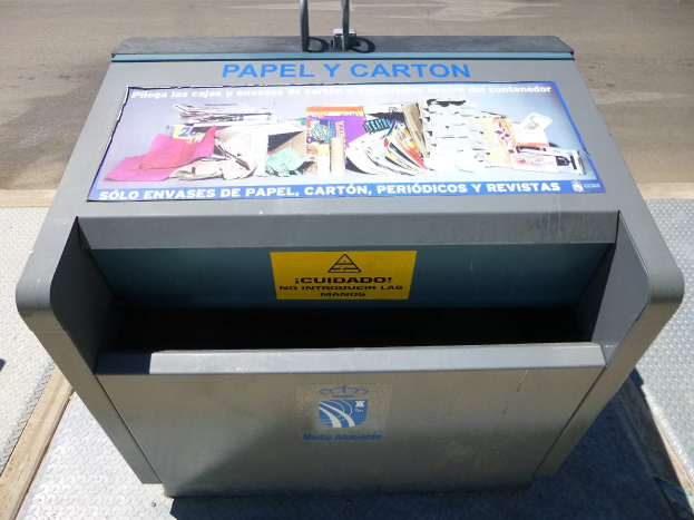 Metallmülltonne am Straßenrand mit einem "Papel y Carton"-Plakat, Fahrzeuge sind im Hintergrund zu sehen.