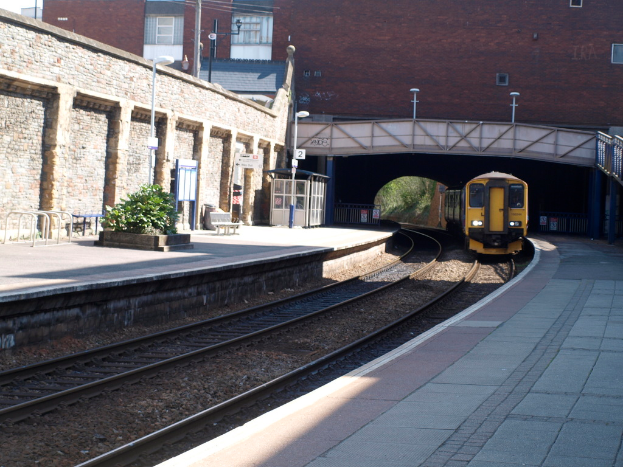 Ein Zug fährt auf Gleisen mit Plattformen auf beiden Seiten, eine Hauswand links, Straßenlaternen, Pflanzen, Bänke, eine Kabine, eine Brücke über die Plattformen und Gebäude und Masten im Hintergrund.