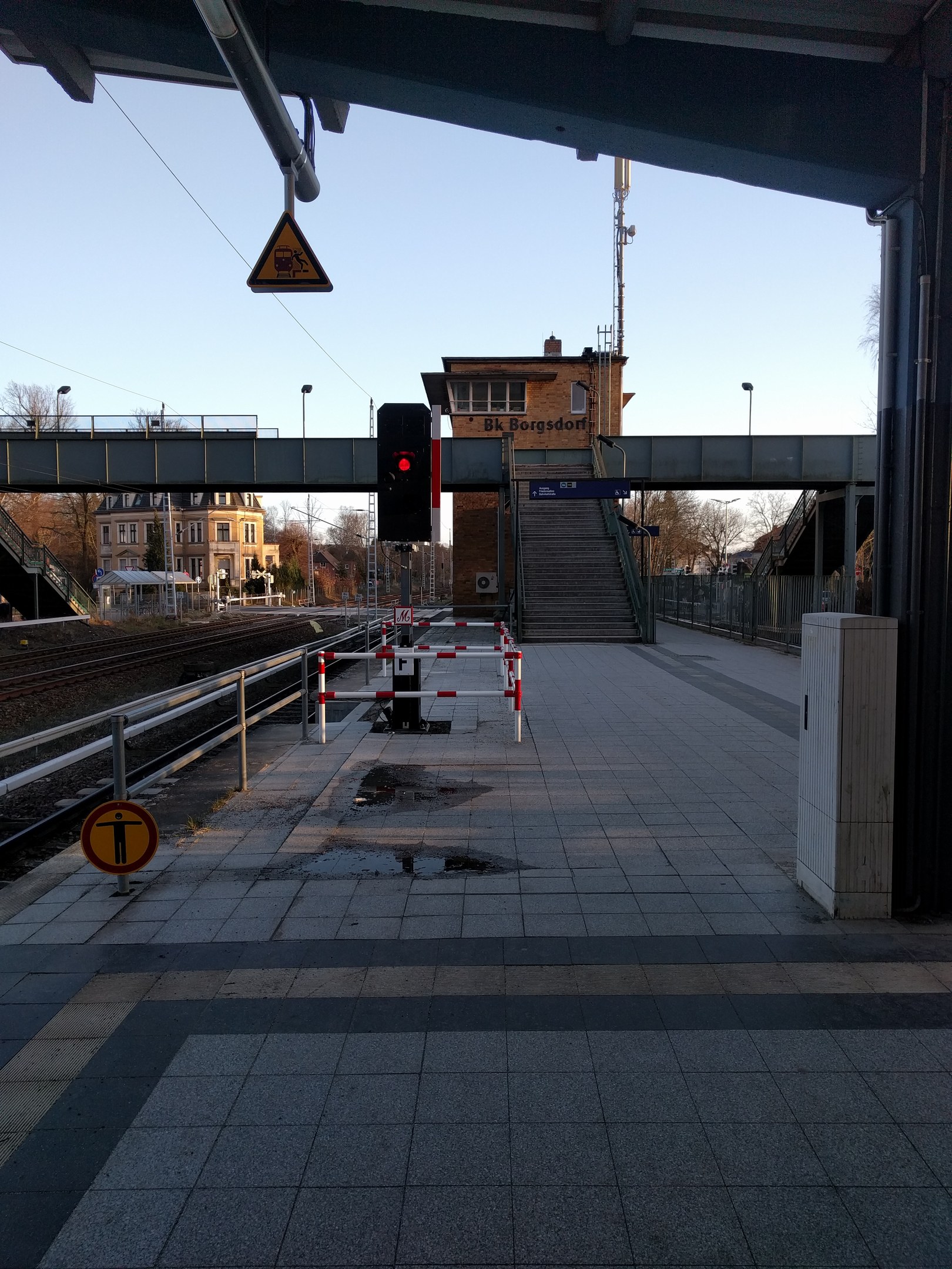 Ein Bahnsteig mit Gleisen, Geländern, einem Schild, einer Ampel an einem Mast, einer Brücke mit Treppen, Laternen, Bäumen, Gebäuden mit Fenstern, einem Turm und einem Himmel im Hintergrund.