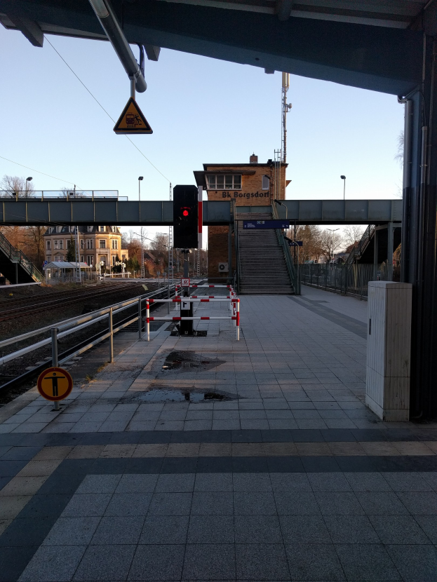 Ein Bahnsteig mit Gleisen, Geländern, einem Schild, einer Ampel an einem Mast, einer Brücke mit Treppen, Laternen, Bäumen, Gebäuden mit Fenstern, einem Turm und einem Himmel im Hintergrund.