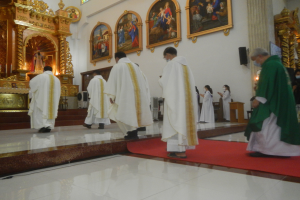 Eine Gruppe von Priestern geht eine rote Teppich in einer Kirche entlang, mit Menschen, die auf dem Boden vor ihnen und auf der rechten Seite stehen, und Foto-Rahmen an der Wand im Hintergrund.