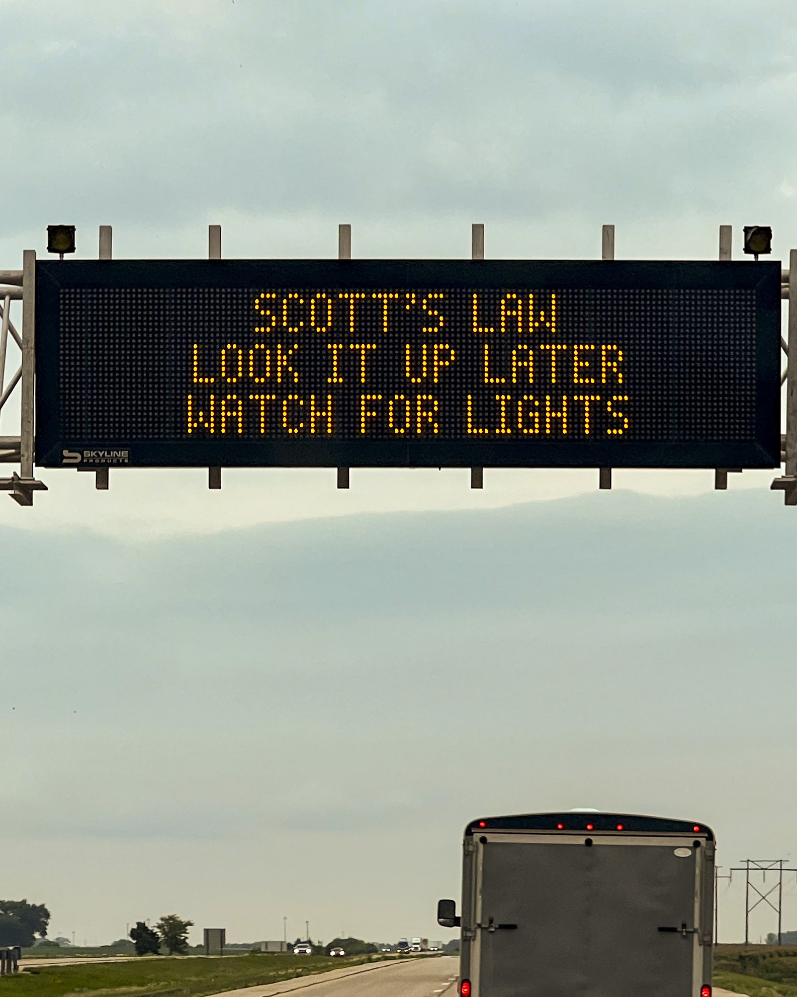 Lkw-Fahrt auf einer Autobahn mit einem "Scott's Law: Schau es später nach, achte auf Lichter"-Schild an der Seite, umgeben von Gras, Bäumen und Strommasten unter einem sichtbaren Himmel.