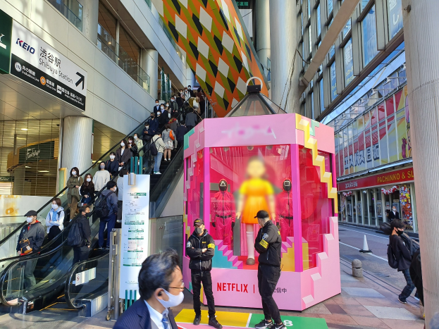 Eine Gruppe von Menschen vor einem Gebäude mit Glaswänden, Säulen und Texttafeln, einige tragen Masken, mit einer Rolltreppe im Hintergrund und einer pinken Box mit der Aufschrift "Netflix's new Netflix Booth in Tokyo".