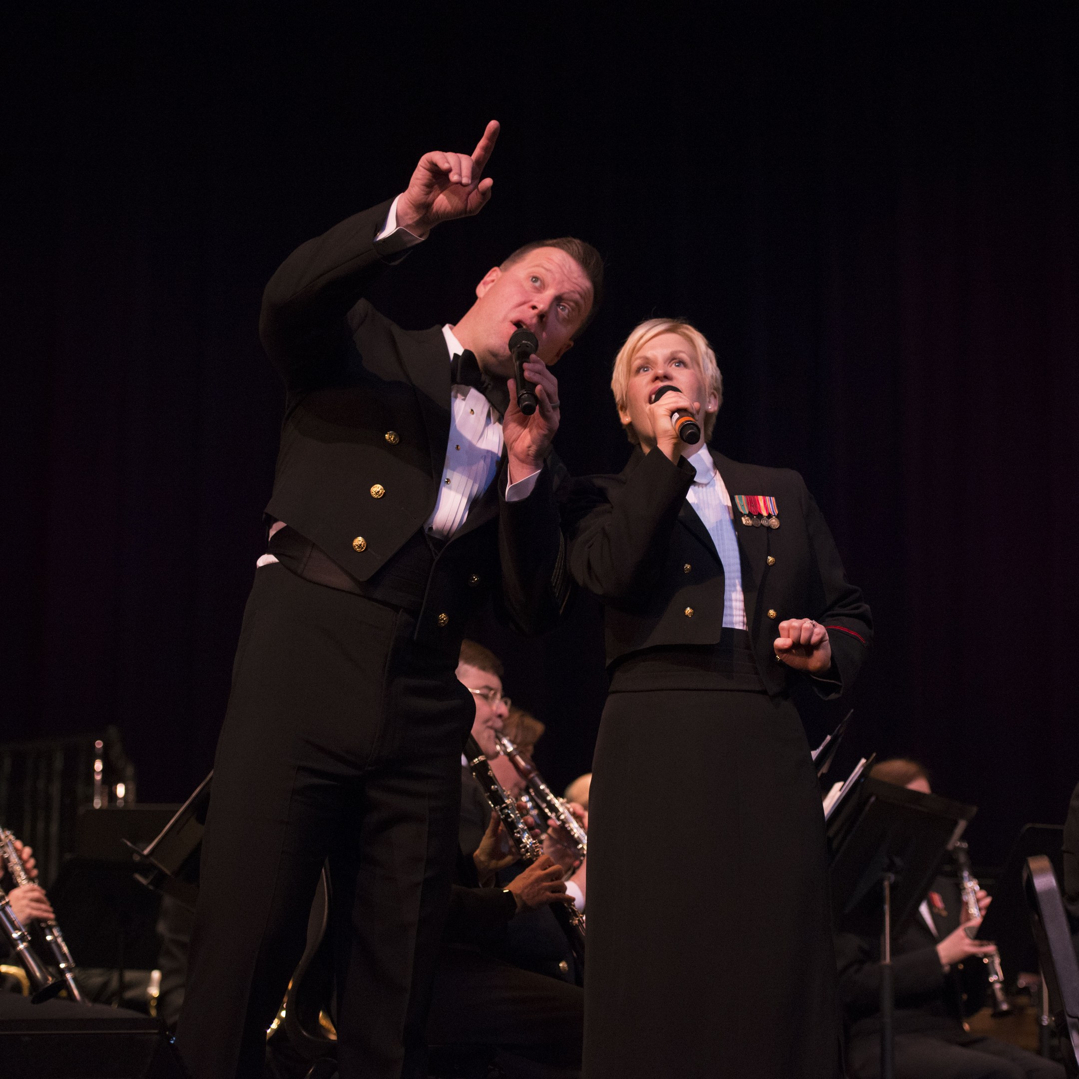 A man and woman in tuxedos holding microphones on a stage, with a band of seated musicians behind them in a dark setting.