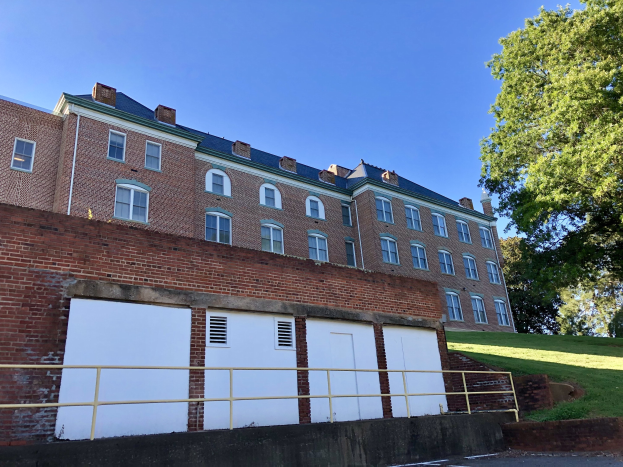 Großes Backsteingebäude mit zahlreichen Fenstern, Metallzaun, Weg, Gras, Bäumen und bewölktem Himmel auf dem Campus der University of Virginia.