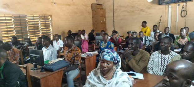 Eine Gruppe von Menschen sitzt an Tischen in einem Klassenzimmer mit Monitoren, Taschen, Büchern und Papieren auf den Tischen, einem Fenster auf der linken Seite und Kartons und einem Fernseher an der Wand im Hintergrund, an einer Techniktrainingssitzung teilnehmend.